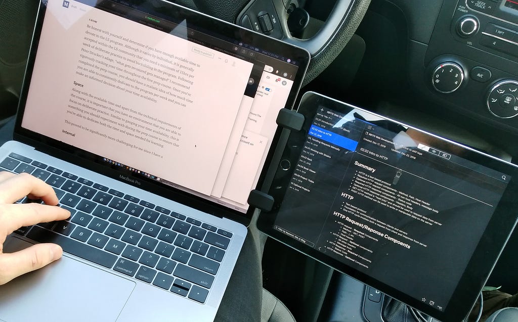 A MacBook with an iPad mounted beside it as a second monitor, set up for studying in the front seat of a car