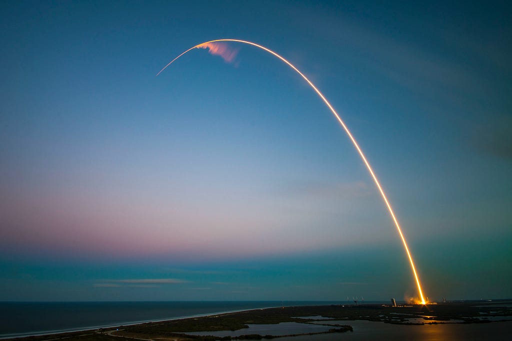 Long-exposure photo of a rocket launch arcing into a twilight sky